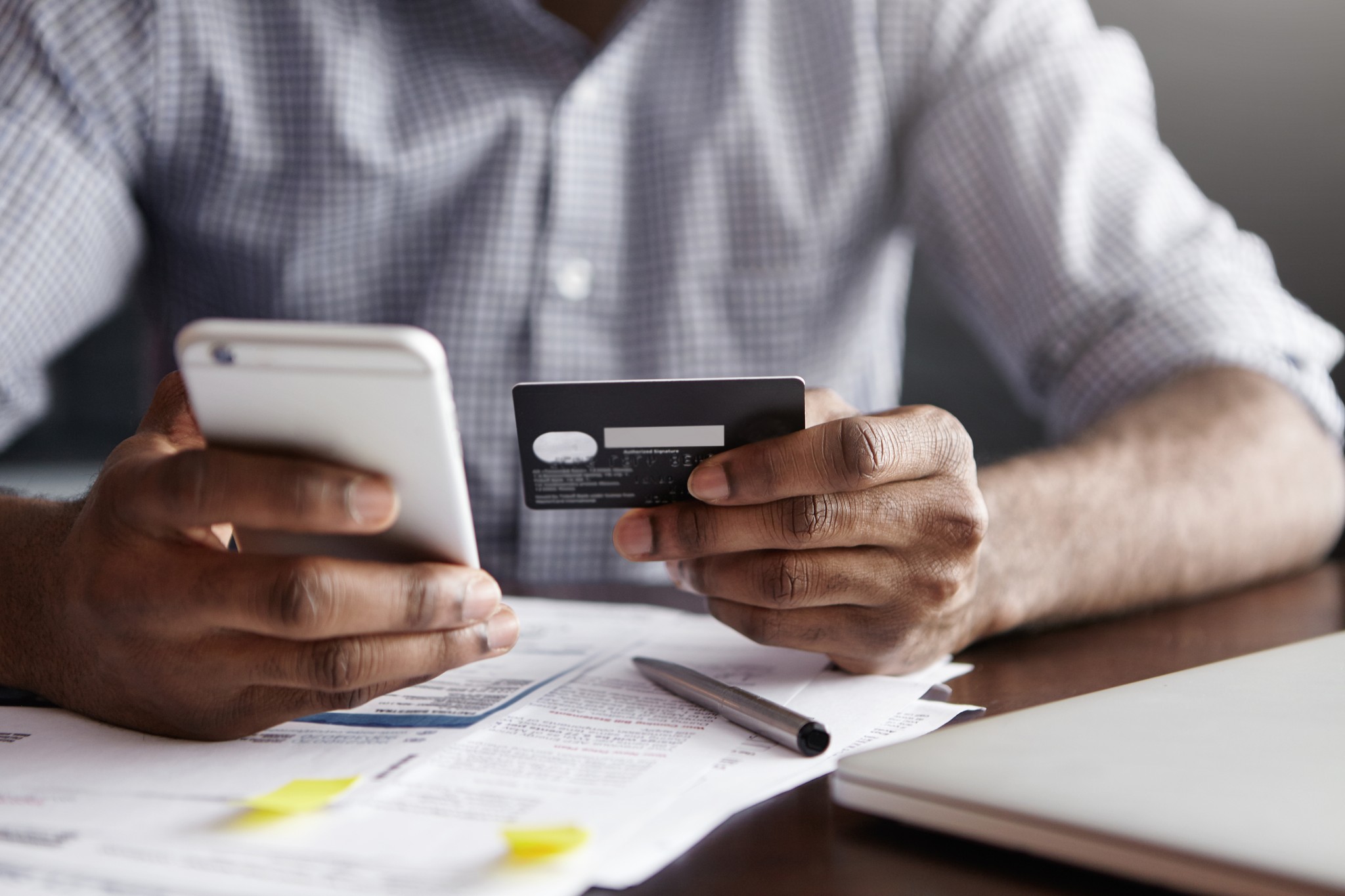Cropped Shot Of African American Male Paying Bill At Restaurant With Online Payment Technology Via Internet, Using Free Wi Fi During Breakfast, Sitting At Table With Plastic Card And Smart Phone